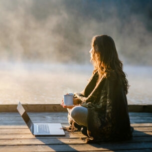 Eine Frau sitzt entspannt mit einer Kaffeetasse vor einem Laptop an einem nebelverhangenen See am Morgen