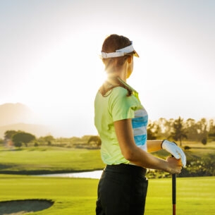 Eine Frau mit Golfschläger in der Hand, die Richtung Sonnenschein über eine weite Rasenfläche schaut