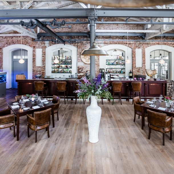 Mehrere eingedeckte Holztische im Restaurants Elements, im Vordergrund eine große weiße Vase mit Blumen, im ein dunkler Bartresen