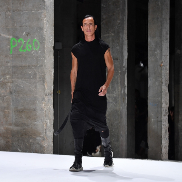 Designer Rick Owens in einem schwarzen, lässigen Kleid läuft in einer Industriehalle über einen Lauftsteg