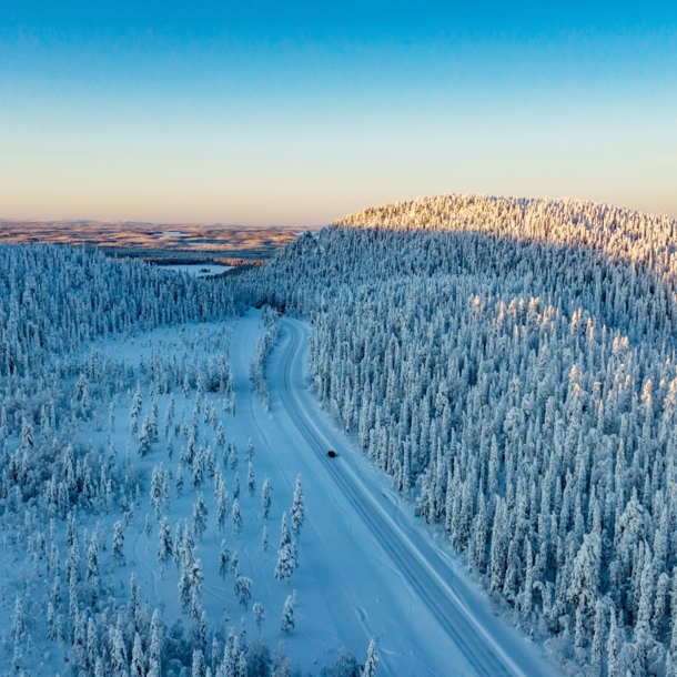 Luftaufnahme einer weiten Winterlandschaft mit schneebedeckten Wäldern, durch die ein einzelnes Auto über eine Straße durch den Schnee in der Abenddämmerung fährt