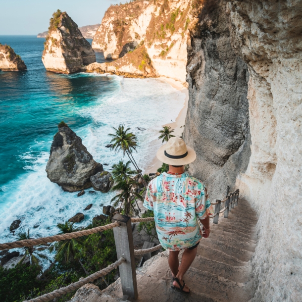 Rückansicht eines Mannes mit Strohhut und Hawaiihemd, der eine schmale Steintreppe an einer Felswand hinunter zu einer palmengesäumten Strandbucht geht