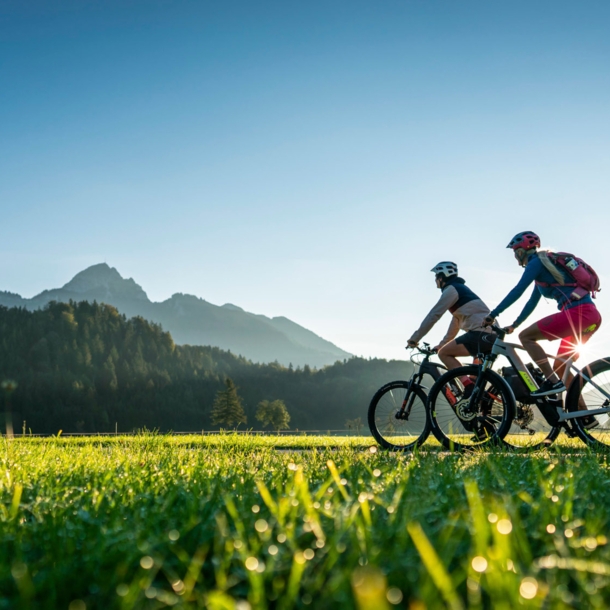 Zwei Personen fahren auf Mountainbikes durch eine Grasebene vor Bergpanorama im Sonnenschein