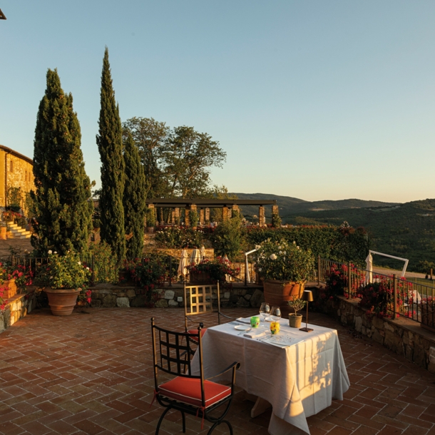 Ein gedeckter Tisch auf einer großen Terrasse einer Villa im toskanischen Baustil mit Blick in eine grüne Hügellandschaft bei Sonnenuntergang