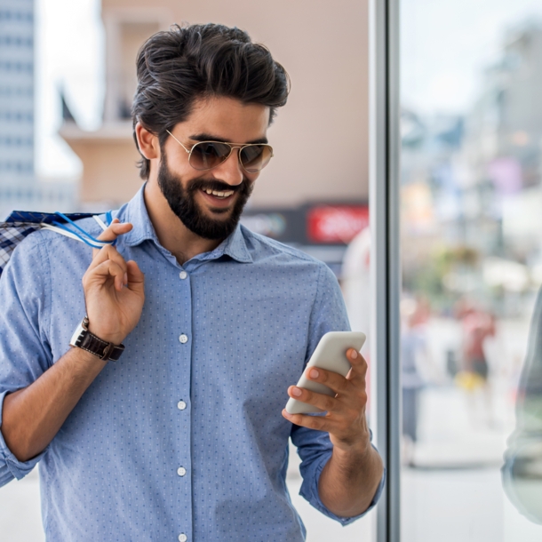 Ein junger Mann mit Vollbart, Sonnenbrille und Einkaufstüten in der Hand schaut in einer Einkaufsstraße lächelnd auf sein Smartphone.