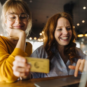 Zwei junge Frauen sitzen lächelnd mit einer Kreditkarte vor einem Laptop in einem Café.