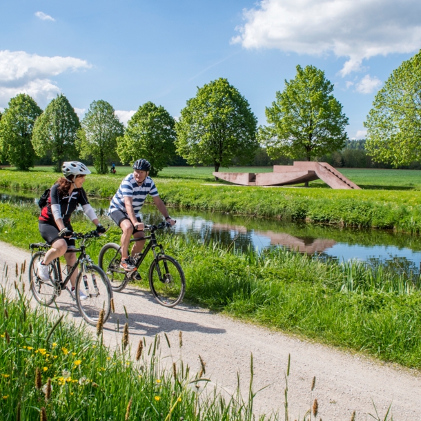 Zwei Personen auf Fahrrädern fahren auf einem Radweg durch eine grüne Landschaft entlang eines Flusses.