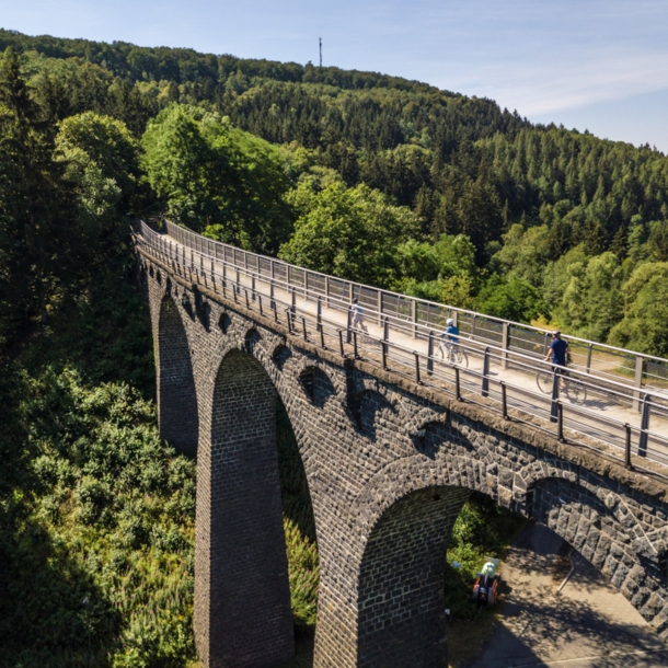 Drei Radfahrende fahren auf einem schmalen Weg auf einem Viadukt in einer hügeligen Waldlandschaft