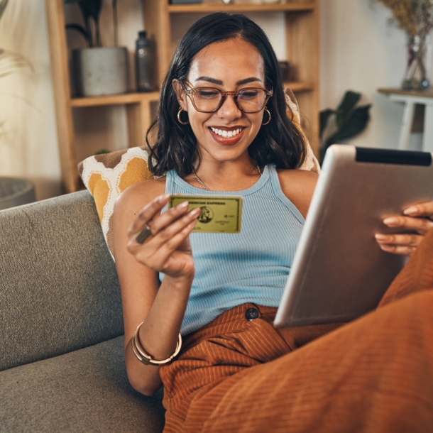 Eine junge Frau sitzt lächelnd auf einem Sofa, in ihren Händen hält sie ein Tablet und eine goldene Kreditkarte von American Express.