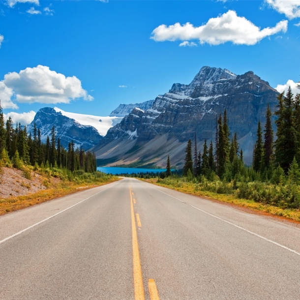 Ein Highway, umringt von Nadelbäumen, im Hintergrund schneebedeckte Berge und ein türkisblauer See.