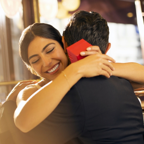 Eine Frau mit einer roten Geschenkbox in den Händen umarmt glücklich lächelnd einen Mann auf einer Sitzbank in einem Restaurant.