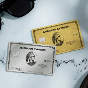 Eine silberne und eine goldene Kreditkarte von American Express liegen auf einem weißen Marmortisch neben einer schwarzen Sonnenbrille, einer Kaffeetasse und einem Teller mit Gebäck.