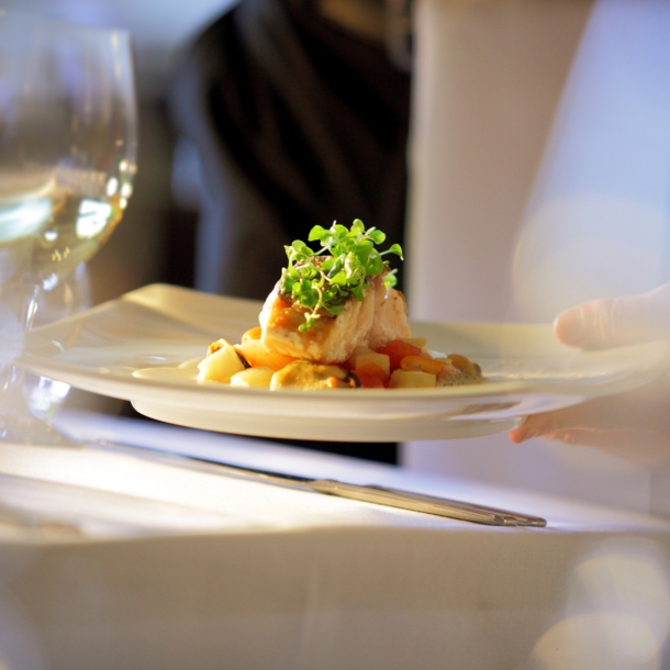 Nahaufnahme eines edel angerichteten Fischgerichtes, das auf einem weißen Teller an einem Tisch mit weißer Tischdecke serviert wird.