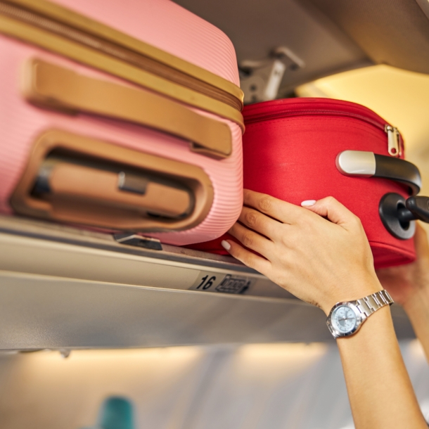 Zwei Hände greifen einen roten Koffer in einem Overhead-Bin eines Flugzeugs, im Vordergrund ein Koffer in Rosa.