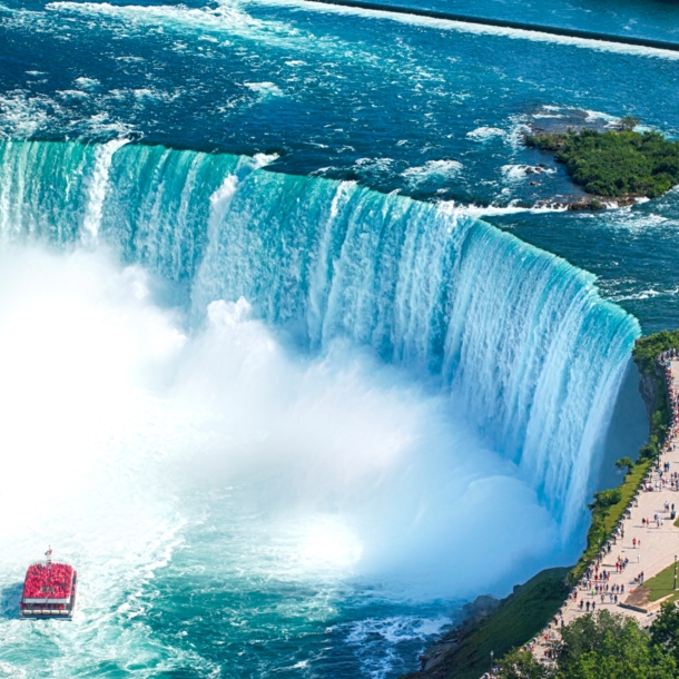 Luftaufnahme eines runden Wasserbeckens mit Boot vor einem Wasserfall, am Ufer eine Aussichtsplattform mit Personen.