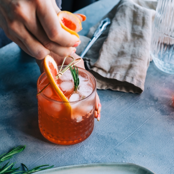 Eine Nahaufnahme einer Person, die einen Cocktail anrichtet, der mit einer Grapefruit-Scheibe und Rosmarin verziert ist.