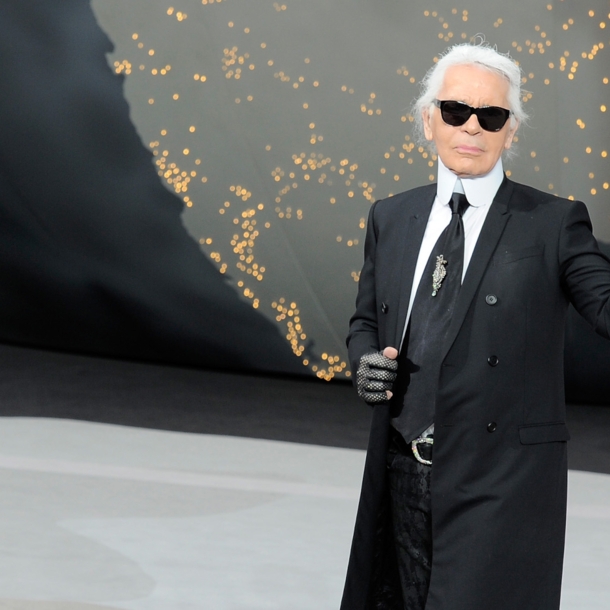 Karl Lagerfeld mit Sonnenbrille im schwarzen Jackett winkt vom Laufsteg einer Modenschau.