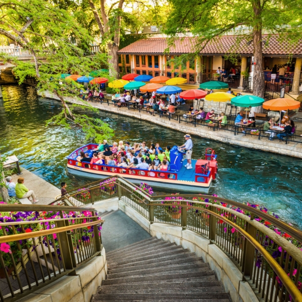 Ein Ausflugsboot fährt auf einem Wasserkanal an einem Restaurant mit Personen auf einer Terrasse mit bunten Sonnenschirmen vorbei, im Vordergrund eine mit Blumen dekorierte Treppe.