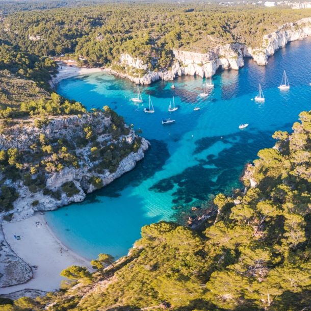 Luftaufnahme eines Küstenabschnitts von Menorca mit Badebuchten, Waldlandschaft und Booten.