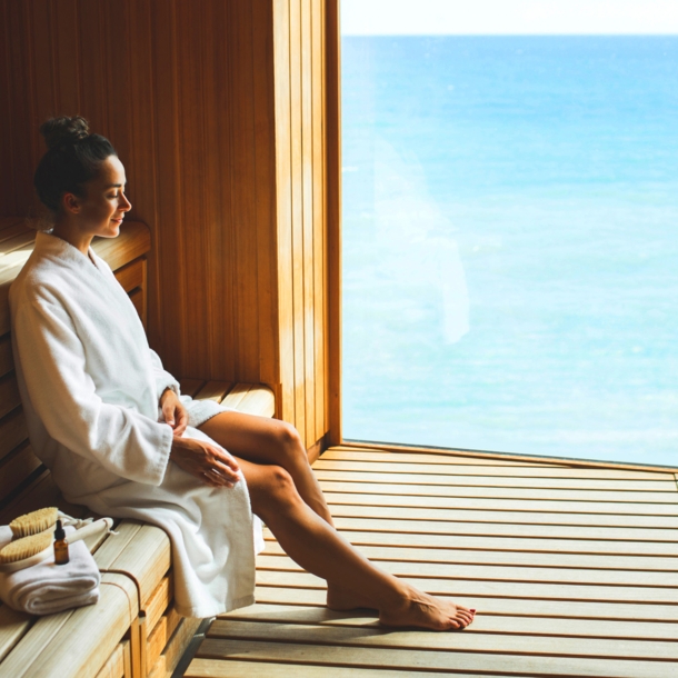 Eine junge Frau im weißen Bademantel sitzt entspannt in einer Holzsauna mit Panoramafenster am Meer.