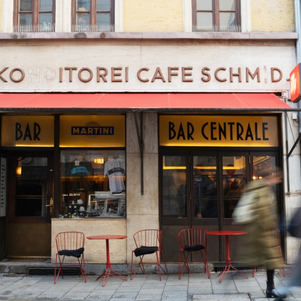 Außenansicht der Bar Centrale mit roter Markise sowie Bistrotischen und Stühlen.