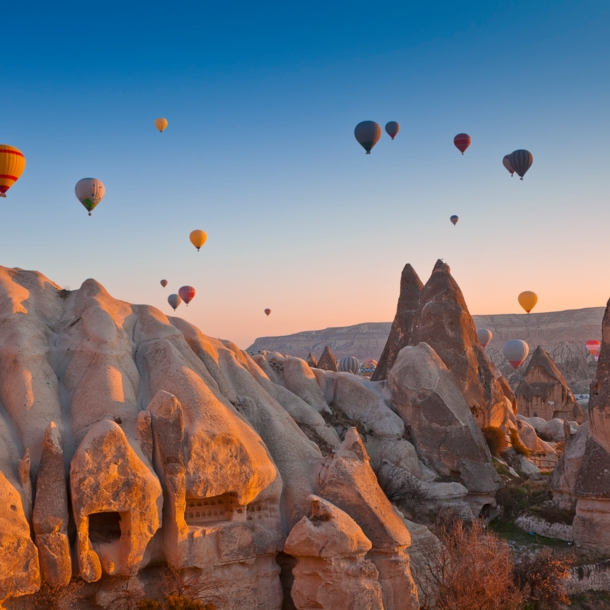 Felsige Landschaft in Kappadokien bei Sonnenuntergang mit zahlreichen schwebenden Heißluftballons im Hintergrund.