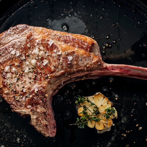 Ein scharf angebratenes Tomahawk-Steak in einer Pfanne. Daneben liegt eine aufgeschnittene Knoblauchknolle, die mit Thymian getoppt ist.