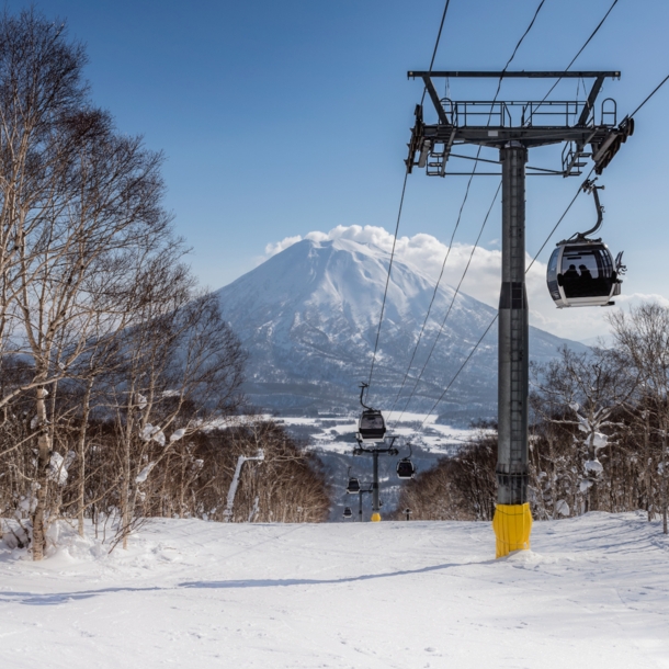 Skilift und Schneelandschaft mit Bäumen. Im Hintergrund ein schneebedeckter Berg und blauer Himmel.