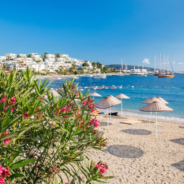 Strand in Bodrum mit Sonnenschirmen mit weißen Häusern an der Küste im Hintergrund.