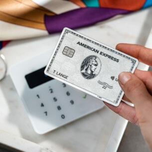 Nahaufnahme einer Hand, die eine silberne American Express Kreditkarte an ein Kartenlesegerät neben Modeaccessoires hält.