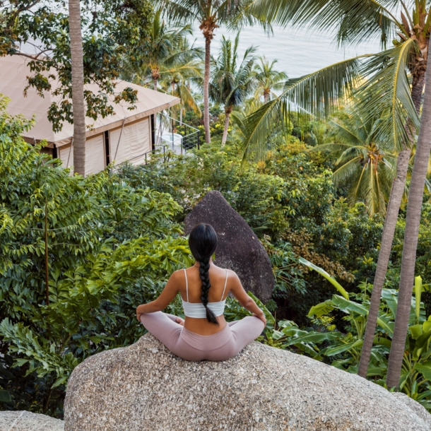 Eine Frau sitzt im Lotussitz auf einem großen Stein, vor ihr eine üppig-grüne Natur, ein Bungalow sowie der Ozean Thailands.