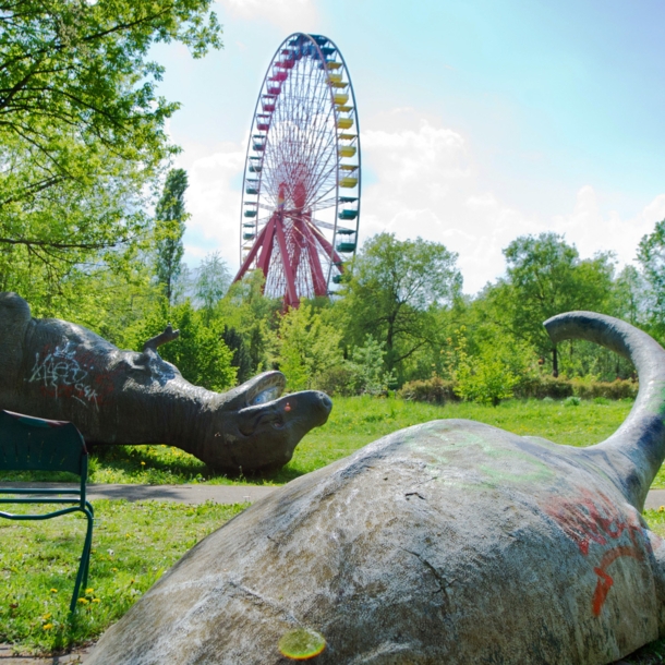 Zwei umgekippte Dinosaurier-Skulpturen auf einer Wiese vor einem Riesenrad in einem Freizeitpark.