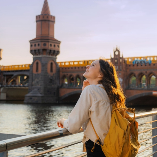 Eine junge Frau mit Rucksack steht am Wasser vor einer Brücke mit Türmen, auf der eine gelbe Straßenbahn fährt.