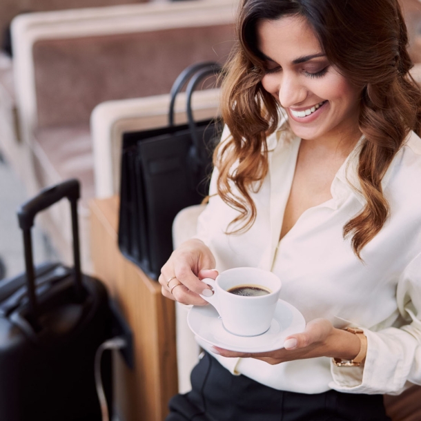 Eine elegant gekleidete Frau sitzt mit einer Tasse Kaffee in einem Sessel, neben ihr steht Gepäck.