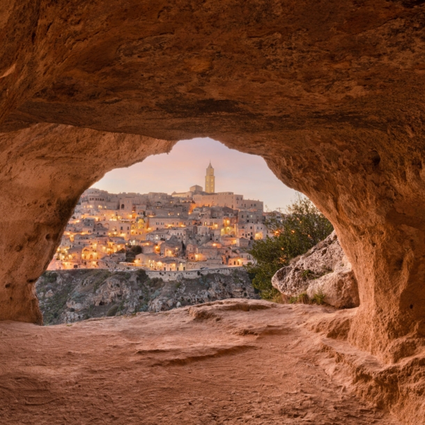 Blick auf die erleuchtete Stadt Matera aus einer Steingrotte im Vordergrund bei Dämmerung.