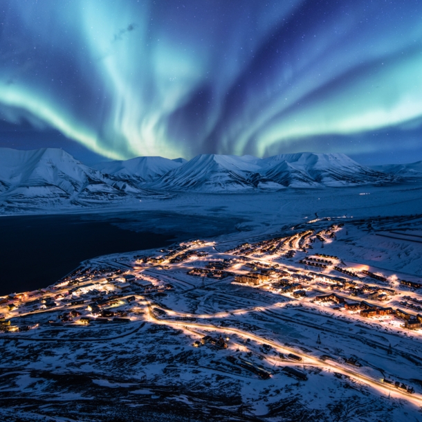 Beleuchtete Ortschaft in schneebedeckter Fjordlandschaft unter Polarlichtern bei Nacht.