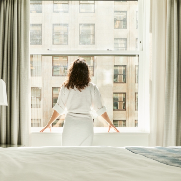 Rückansicht einer Frau im Bademantel, die am Fenster eines Hotelzimmers steht.