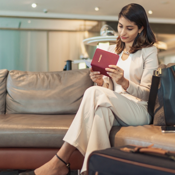 Eine Frau sitzt mit überschlagenen Beinen auf einem hellen Ledersofa und sieht sich Reiseunterlagen an.