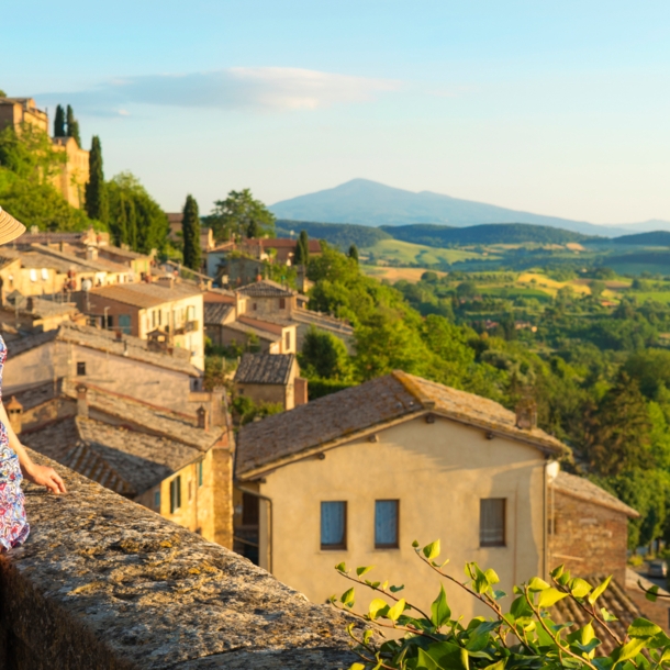 Eine Frau im Sommerkleid blickt von einer Mauer in einem italienischen Dorf hinunter auf eine grüne Hügellandschaft bei warmem Sonnenlicht.