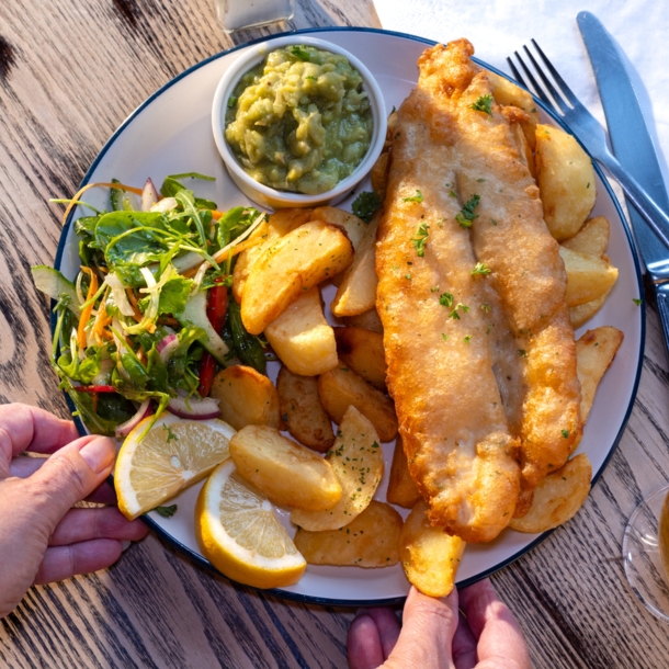 Eine Hand greift nach einem Teller Fish and Chips mit Beilagen auf einem Holztisch bei Sonnenschein.