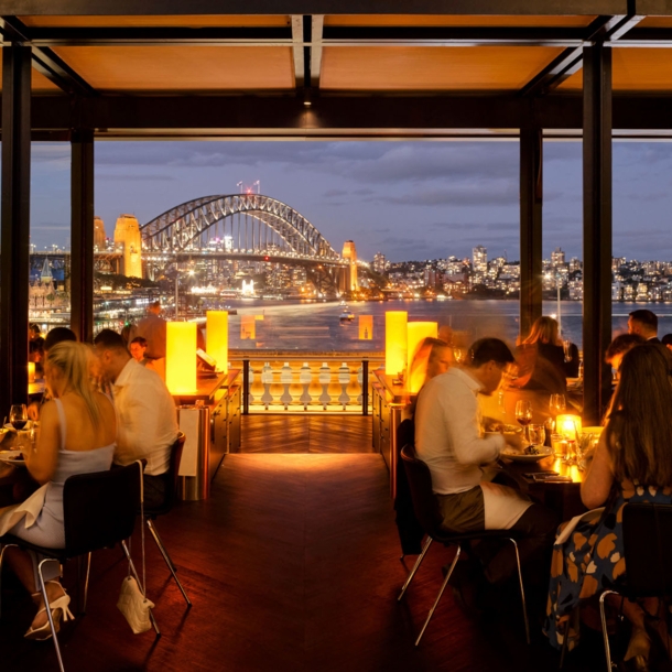 Außengastronomie am Hafen von Sydney vor beleuchteter Harbour Bridge bei Nacht.