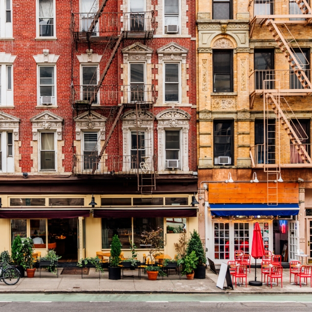 Geschlossene New Yorker Häuserfront mit Feuerleitern und Restaurants mit Außengastronomie.