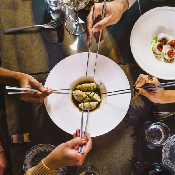 Aufsicht eines gedeckten Restauranttisches mit einem weißen Teller mit Teigtaschen, umgeben von vier Händen mit Essstäbchen.