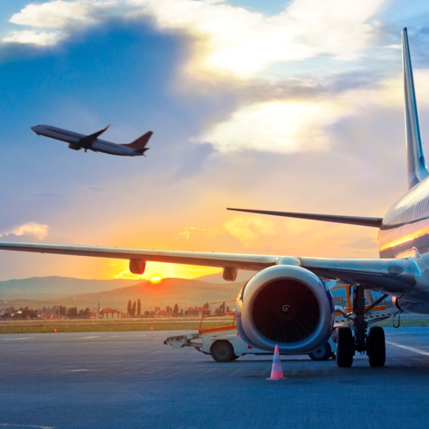 Ein Flugzeug am Himmel über einem geparkten Flugzeug am Flughafen vor einer hügeligen Landschaft bei Sonnenuntergang.