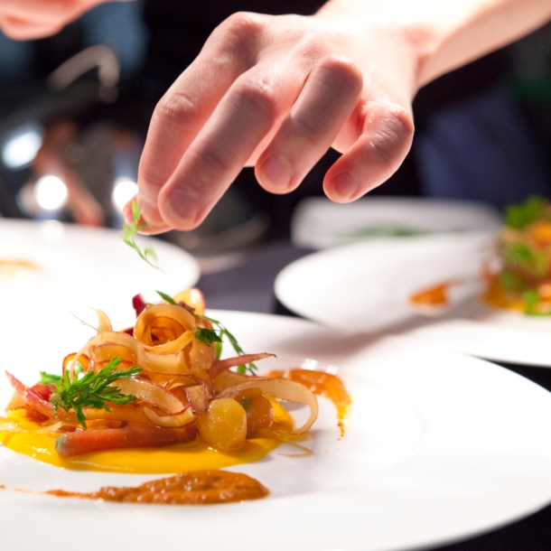 Eine Hand garniert ein Gericht auf einem weißen Teller in einer Restaurantküche.