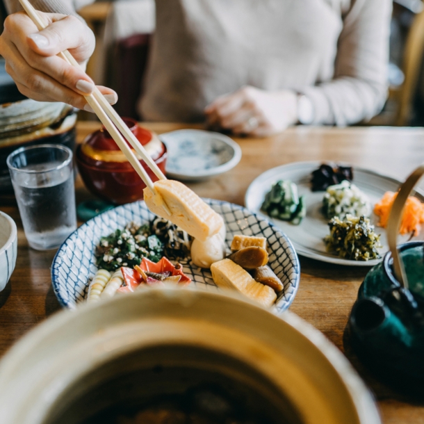 Auf einem Tisch stehen mehrere Schälchen mit verschiedenen asiatischen Gerichten. Eine unkenntliche Person hält mit Stäbchen etwas zu essen hoch.