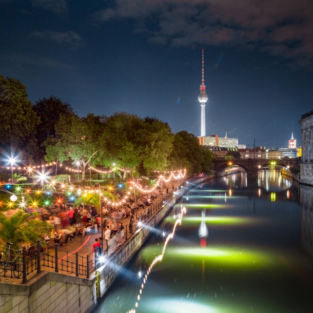 Personen in einer Strandbar am Spreeufer an der Berliner Museumsinsel bei Nacht, im Hintergrund ein beleuchteter Fernsehturm.