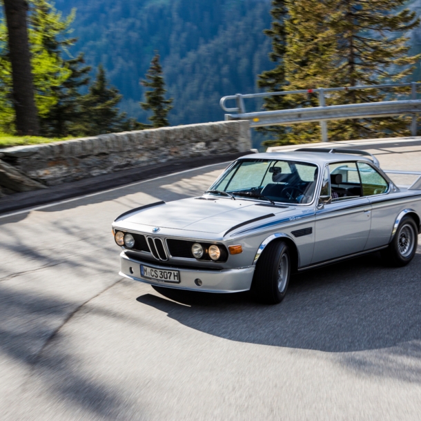 Ein fahrender silberner BMW 3.0 CSL in einer Kurve auf einer Straße in den Bergen.