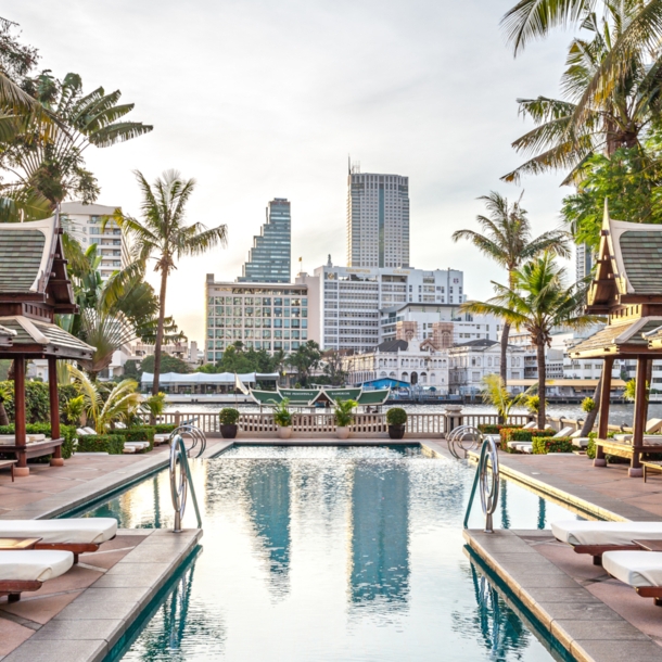 Eine mit Palmen gesäumte Poolanlage im Hotel The Peninsula in Bangkok. Am Wasser stehen Holzliegen mit weißem Bezug, einige befinden sich unter Pagoden-Dächern.