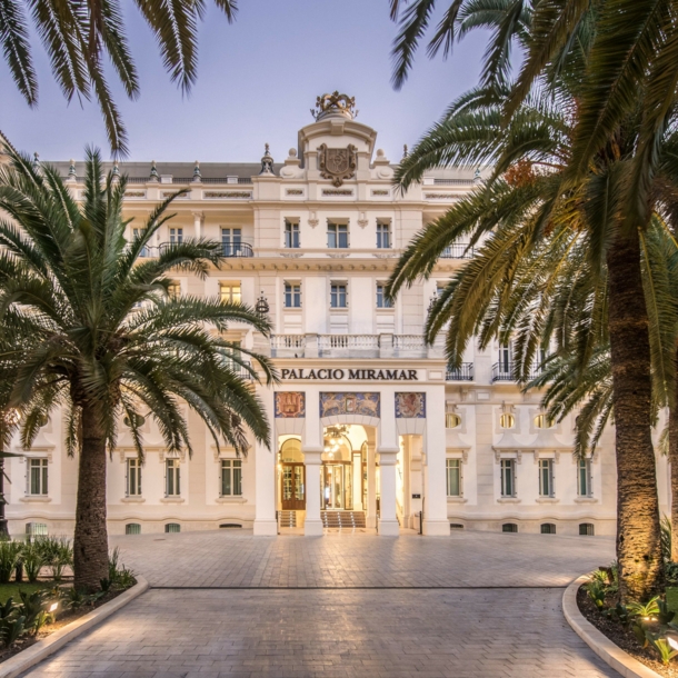 Imposante, helle Frontfassade eines Grandhotels mit Vorplatz, im Vordergrund Begrünung mit Palmen.